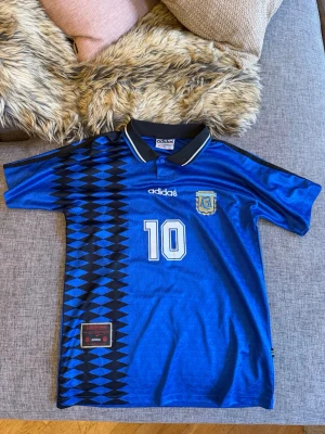 Blå Argentina tröja Adidas Maradona - Säljer en blå Argentina fotbollströja från Adidas med nummer 10 och Maradona på ryggen. Tröjan har svartvitt mönster på sidan, klassisk krage och broderad Argentina-logga på bröstet. Perfekt för dig som älskar retro och fotboll.