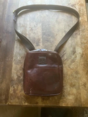 Brun axelväska i läder - Snygg brun axelväska i äkta läder med justerbar axelrem i tyg. Väskan har en fyrkantig form och ett diskret emblem på framsidan. Perfekt för dig som vill ha en stilren och klassisk look med plats för det viktigaste.