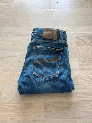 Blå jeans från Nudie Jeans - Säljer ett par klassiska blå jeans från Nudie Jeans med smal passform. Jeansen har fem fickor, orangea sömmar och Nudie-loggan på bakfickan. Perfekta till en avslappnad stil och tillverkade i slitstarkt denimtyg. W28 L32