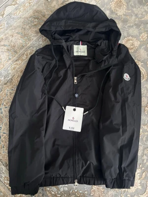 Svart vindjacka från Moncler - Snygg svart vindjacka från Moncler med huva och dragkedja framtill. Jackan har elastiska muddar vid ärmslut och nederkant samt Moncler-logga på ärmen. Tillverkad i lätt och tåligt material, perfekt för blåsiga dagar.