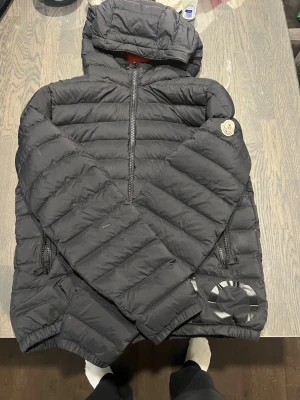 Moncler Svart dunjacka med huva - Snygg svart dunjacka med huva och två dragkedjeförsedda fickor. Jackan har ett emblem på ärmen och stora bokstäver 'MO' tryckta nedtill. Puffig modell med horisontella sömmar och elastiska muddar vid ärmslut och nederkant. Perfekt för kalla vinterdagar.