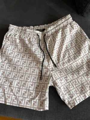 Vita Fndi badshorts monogram - Snygga badbyxor med all-over logomönster i beige. Shortsen har elastisk midja med svart snörning och sidofickor samt en bakficka. Perfekta för sommaren och ger en lyxig vibe till din strandoutfit! Storlek L men lite mindre i storleken!