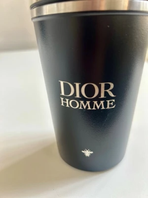 Dior Homme termosmugg - Exklusiv termosmugg från Dior Homme med silverfärgad kant upptill och praktiskt lock.Metall invändigt som gör att drycken håller värmen. Muggen har en stilren design med tydlig Dior Homme-logga på framsidan. Äkta GWP från Dior. Aldrig använd helt ny. Perfekt för morgonkaffet när du är på väg till skolan eller jobbet! 