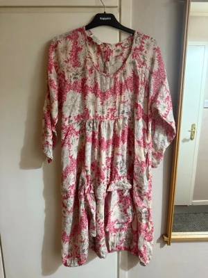 Blommig röd och vit klänning - Luftig klänning med blommigt mönster i rött och vitt. Modellen har trekvartsärm, rund halsringning och knappar i ryggen. Passformen är loose och tyget känns mjukt, troligen bomull. Perfekt för varma dagar och enkel att styla.