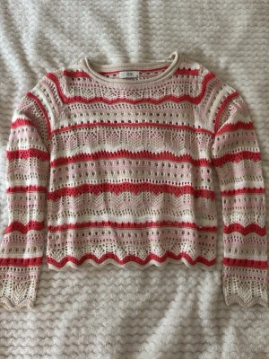 Stickad tröja i rosa och rött från JDY - Söt stickad tröja från JDY i storlek XS. Tröjan har ett genombrutet mönster i vitt, rosa och rött med vågig kant nertill och långa ärmar. Perfekt för dig som gillar en feminin och trendig look. Materialet känns mjukt och luftigt. Inte använt