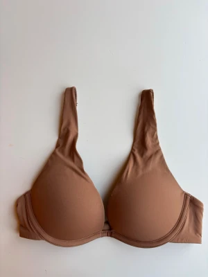 Nude vadderad  T-shirt bh med bygel storlek 75b från H&M - Säljer en slät, nude-färgad T-shirt bh med bygel och vadderade kupor. Bh:n har breda axelband och hakknäppning bak. Materialet är mjukt och stretchigt, perfekt under åtsittande plagg för en osynlig look. Storlek 75b från H&M. Använd två gånger.