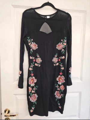 Klänning mesh H&M Divided 40 - Svart långärmad klänning från Divided med mesh upptill och broderade blommor i rosa, rött och grönt längs framsidan. Klänningen har en kroppsnära passform och transparenta ärmar som ger en cool kontrast till de färgglada broderierna.  Kan även hämtas I Partille  E4