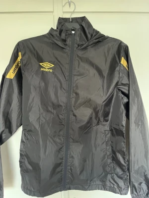 Svart vindjacka från Umbro med huva - Snygg svart vindjacka från Umbro med gula loggor på bröstet, ärmen och ryggen. Jackan har dragkedja framtill, huva och elastiska muddar vid ärmsluten. Tillverkad i lätt och glansigt material som skyddar mot vind.