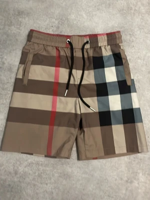 Rutig shorts med snörning - Säljer ett par snygga shorts med klassiskt rutigt mönster i beige, brunt, svart, vitt och rött. Shortsen har elastisk midja med svart snörning och sidofickor. Perfekta för sommaren och ger en trendig look.