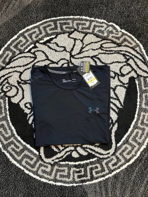 Svart tränings-tshirt från Under Armour - Snygg svart tränings-tshirt från Under Armour med diskret logga på bröstet och tryckt text längs sidan. Tillverkad i ett lätt och snabbtorkande funktionsmaterial som passar perfekt till gymmet eller löprundan. Klassisk rund hals och korta ärmar.