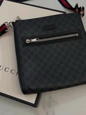 Svart axelväska från Gucci - Snygg svart axelväska från Gucci med diskret GG-mönster över hela väskan. Väskan har en silverfärgad dragkedja på framsidan och en justerbar axelrem i blått och rött. Materialet är läder och detaljerna är i metall.