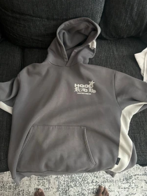 Grå hoodie med vit text och ficka - Snygg grå hoodie med vit text och grafiskt tryck på bröstet. Hoodien har en stor magficka och vita detaljer längs sidorna. Perfekt för en avslappnad streetwear-look. Mjuk bomullskänsla och klassisk huva.