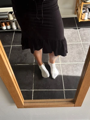 Svart volangkjol med midjeresår - Säljer en svart kjol med volangkant nedtill och resår i midjan. Kjolen har en figurnära passform och slutar strax under knät. Perfekt att styla med en enkel topp för en trendig look.