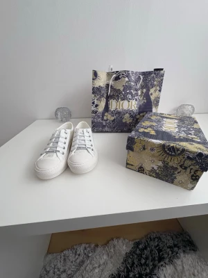 Vita Dior sneakers med logga - Snygga vita låga sneakers från Christian Dior med tydlig logga på snörningen. Skorna har klassisk siluett, vita snören och är tillverkade i textil med gummisula. Perfekta för dig som vill ha en clean och exklusiv look.