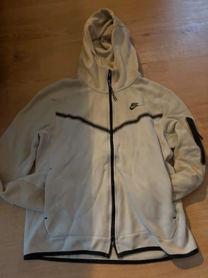 Beige Nike hoodie med dragkedja - Snygg beige hoodie från Nike med hel dragkedja och svart logga på bröstet. Hoodien har svarta detaljer längs dragkedjan, muddar och en rand över bröstet. Praktisk ficka på ärmen och luva för extra stilpoäng. Perfekt för en avslappnad streetwear-look.