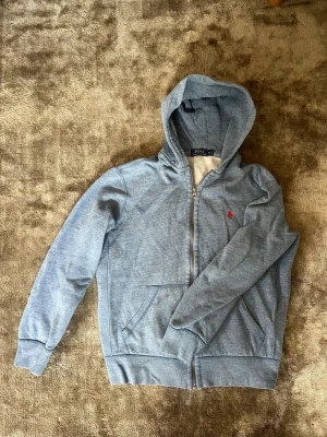 Blå hoodie från Polo Ralph Lauren - Snygg ljusblå hoodie från Polo Ralph Lauren med dragkedja och huva. Klassisk röd logga broderad på bröstet och mjukt, vitt foder på insidan. Två fickor framtill och ribbade muddar vid ärmar och nederkant. Riktigt schysst passform. Kan även passa S .Om du har frågar är det bara att fråga på. Pris går att diskutera med seriösa köpare. Kolla in min profil och se om du hittar något mer intressant. Mvh Larsen_Closet