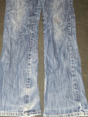Blå bootcut jeans med slitna detaljer - Ett par klassiska blå bootcut jeans med slitna detaljer och lätt tvättad look. Jeansen har vida benslut och synliga slitningar vid fållen för en avslappnad stil. Perfekta för dig som gillar en retro vibe och vill ha ett par jeans med karaktär.