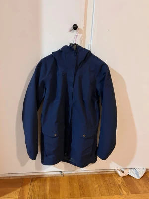Norrøna jacka - Snygg mörkblå vindjacka med dragkedja framtill och två stora fickor med knappar. Jackan har en praktisk huva och är tillverkad i ett lätt, vattenavvisande material. Perfekt för blåsiga dagar och enkel att matcha med olika outfits. Använd ganska lite och är i storlek S