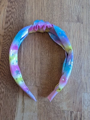 Färgglatt diadem med stjärnmönster - Säljer ett diadem i tyg med regnbågsfärger i rosa, blått, gult och lila. Diademet har ett knutliknande parti upptill och är dekorerat med små vita prickar och silverfärgade stjärnor. Materialet är mjukt och elastiskt, perfekt för att piffa upp din look.