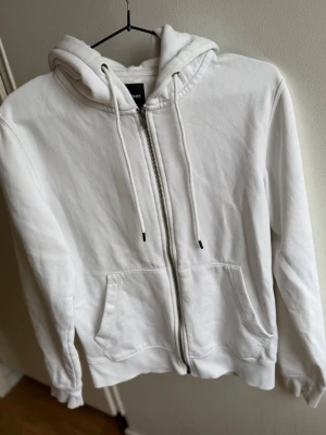 Vit zip hoodie med huva från Jack & Jones  - Klassisk vit hoodie med dragkedja framtill och två stora fickor. Tröjan har justerbar huva med snören och ribbade muddar vid ärmar och nederkant. Perfekt för en avslappnad och clean look.