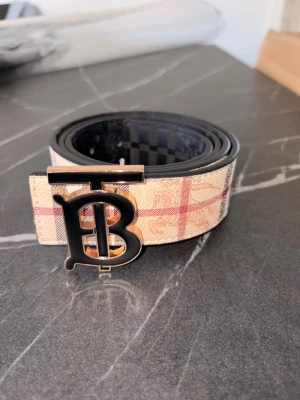 Beige bälte med rutigt mönster - Snyggt bälte i beige med klassiskt rutigt mönster och stor metallspänne med bokstäver. Bältet har svarta detaljer och känns riktigt trendigt. Perfekt accessoar för att lyfta din outfit.