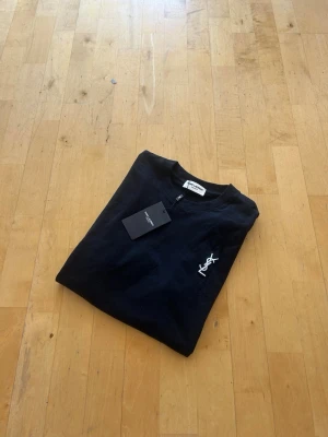 Svart t-shirt från Saint Laurent - Snygg svart t-shirt från Saint Laurent med klassisk rund hals och vit YSL-logga broderad på bröstet. Tillverkad i mjuk bomull för en bekväm känsla. Perfekt för dig som gillar stilrena och exklusiva plagg.