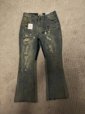 Gallery dept jeans  - Unika grå jeans med bootcut passform och rå, avklippt kant nertill. Jeansen har slitningar, färgstänk och patch-detaljer framtill för en edgy look. Klassisk femficksmodell i denim med låg midja. Perfekta för dig som gillar streetstyle och statement-plagg.