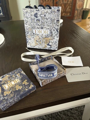 Blått armband från Christian Dior - Säljer ett blått tygarmband från Christian Dior med texten 'CHRISTIAN DIOR J'ADIOR' broderat i vitt. Armbandet har justerbar knytning med tofsar i ändarna och levereras i en exklusiv Dior-presentförpackning.