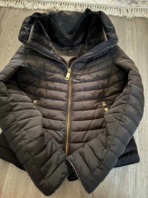 Svart pufferjacka med huva - Säljer en svart pufferjacka med guldfärgade dragkedjor och mysigt foder i huvan. Jackan har två fickor med dragkedja och quiltad design som ger en schysst look. Perfekt för kalla dagar när du vill hålla dig varm och snygg.