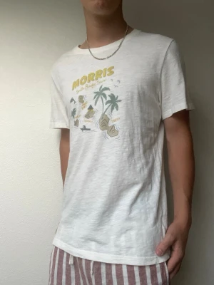 Morris T-shirt med tryck - vit - Strl M - Snygg vit T-shirt från Morris med somrigt tryck framtill. Perfekt till vardags eller semestern. Mjuk och skön passform.  • Bra skick • Normal passform • Modellen på bilden är 178 cm / 70 kg för referens.