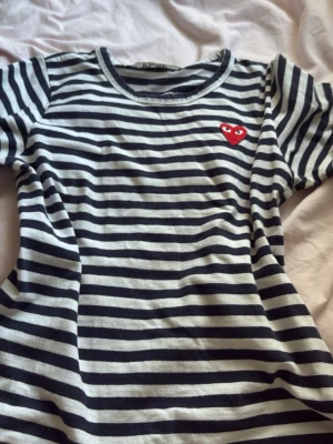 Randig t-shirt med rött hjärta - Säljer en svartvit randig t-shirt med ett broderat rött hjärta på bröstet. T-shirten har rund halsringning och korta ärmar. Perfekt för dig som gillar klassisk stil med en twist.