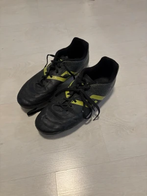 Svarta fotbollsskor Pro Touch - Säljer ett par svarta fotbollsskor från Pro Touch med gula detaljer och snörning. Skorna har en klassisk form med rundad tå och mönstrad ovandel. Perfekta för dig som vill ha bra grepp på planen och snygg stil.