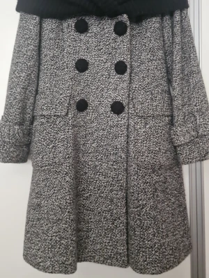 Gråmelerad kappa med stor krage Etam 32% wool 165/84A - Snygg gråmelerad kappa med dubbelknäppning och stora svarta knappar. Kappan har en bred, ribbstickad svart krage och uppvikta ärmslut. A-linjeformad passform som ger en trendig siluett. Perfekt för kyliga dagar och ger en stilren look.