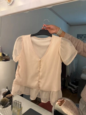 Ljusrosa blus med volang och puffärm - Supersöt ljusrosa blus med transparenta puffärmar och volangkant nedtill. Blusen har v-ringning och knäppning framtill. Tillverkad i ett tunt, lätt material som ger en romantisk vibe. Perfekt att styla med jeans eller kjol för en trendig look.