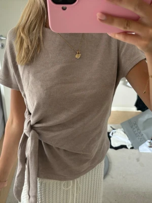 Beige ribbad topp med knyt - Säljer en beige ribbad topp med korta ärmar och snygg knytning i sidan. Toppen har en rund halsringning och är gjord i ett mjukt material som känns skönt mot huden. Perfekt att styla med både jeans och kjol.