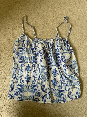 Vit och blå mönstrad topp Primark -  luftig vit blus  från Primark med blått blommigt  mönster💕. Toppen har tunna axelband och en avslappnad passform. Missfärgning på baksidan av toppen. Passar mig som har storlek S