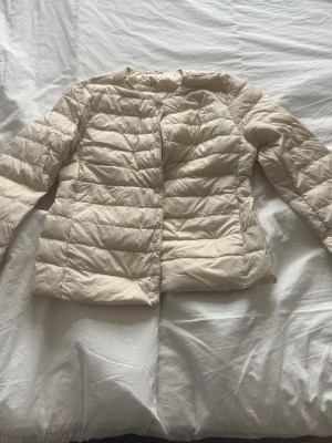 Beige quiltad dunjacka - Snygg beige dunjacka med quiltad design och lång ärm. Jackan har en enkel, stilren look utan synliga dragkedjor eller knappar framtill. Perfekt för kyligare dagar och lätt att matcha med olika outfits.