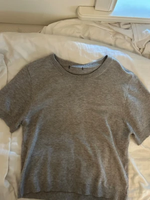 Grå stickad kortärmad topp - En enkel och stilren grå stickad topp med korta ärmar och rund hals. Perfekt att matcha med jeans eller kjol för en clean look. Mjuk och skön kvalitet med ribbade kanter vid hals, ärmslut och nederkant.