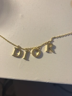 Guldhalsband med DIOR-bokstäver - Snyggt halsband i guldfärg med hängande bokstäver som stavar DIOR. Bokstäverna är i metall och O:et är dekorerat med små pärlor för extra bling. Perfekt accessoar för dig som gillar statement-smycken och exklusiv känsla.