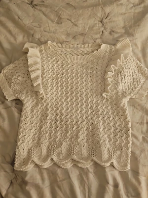 Vit stickad topp med volangdetaljer - Söt vit stickad topp med korta ärmar och volangdetaljer på axlarna. Toppen har ett fint hålmönster och vågig kant nedtill. Perfekt för en trendig och feminin look. Materialet känns mjukt och luftigt.