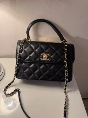 Svart quiltad handväska från - Snygg svart handväska från  i quiltat läder med guldfärgade detaljer och kedjerem. Klassisk modell med topphandtag och ikonisk -logga framtill. Perfekt accessoar för dig som vill ha en tidlös och lyxig look.