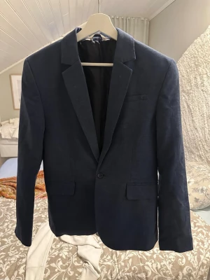 Mörkblå klassisk kavaj - Stilren mörkblå kavaj med två framfickor och en bröstficka. Kavajen har smal passform, enkel knappstängning och klassiska slag. Perfekt för en dressad look och funkar till skjorta eller t-shirt. Materialet ser ut att vara en mjuk bomullsblandning.