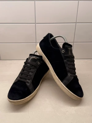 Svarta Saint Laurent sneakers  - Snygga svarta sneakers från Saint Laurent i mocka med detaljer i läder och svarta skosnören. Klassisk låg modell med vit sula och diskret logga på plösen. Perfekta för en stilren och trendig look. Enda defekten är att de är några små jack vid anklarna (kika på bilderna) 