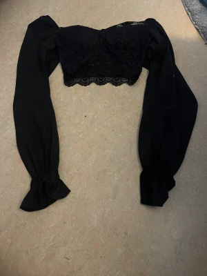 Svart croppad spetsblus med puffärm - Supersnygg svart croppad blus med långa puffärmar och spetsdetaljer. Toppen har en vacker v-ringning och spetskant nedtill som ger en romantisk vibe. Perfekt att styla med höga jeans eller kjol för en trendig look.