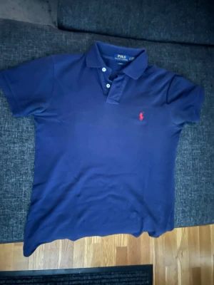 Marinblå pikétröja från Ralph Lauren - Klassisk marinblå pikétröja från Ralph Lauren med röd broderad logga på bröstet. Tröjan har korta ärmar, krage och knappar framtill. Perfekt för en stilren och avslappnad look.