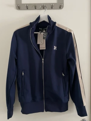Palm angels fullzip - Snygg mörkblå track jacket från Palm Angels med vita ränder längs ärmarna och dragkedja framtill. Jackan har ståkrage, två fickor med dragkedja och logga på bröstet. Perfekt för en sportig och trendig look.