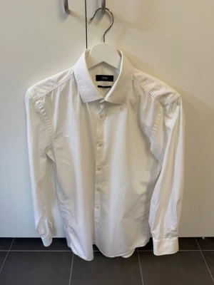 Vit skjorta från Hugo boss, stl 38 - Klassisk vit skjorta från Hugo boss i slim fit-modell. Skjortan har långa ärmar, knappar framtill och en stilren krage. Perfekt för dig som gillar en clean och tidlös look.