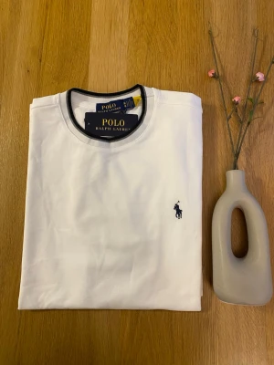 Vit t-shirt från Polo Ralph Lauren - Klassisk vit t-shirt från Polo Ralph Lauren med svart logga på bröstet och svarta detaljer vid krage och ärmslut. T-shirten har rund hals och är tillverkad i mjuk bomull för en skön känsla. Perfekt till jeans eller shorts.