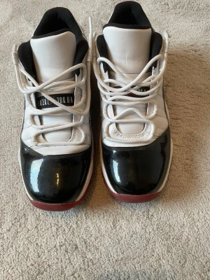 Svarta och vita låga sneakers - Säljer ett par snygga Air Jordan 11 Retro Low “Concord Bred” i fint begagnat skick. Klassisk färgkombination med vit ovandel, svart lackläder och röd yttersula som passar till det mesta.