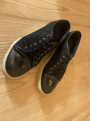 Svarta låga sneakers med gummitå - Säljer ett par svarta sneakers med lågt skaft och vita sulor. Skorna har svarta skosnören, metallöljetter och en blank gummitå som ger en cool kontrast till den matta ovandelen. Perfekta för dig som gillar en stilren och enkel look.